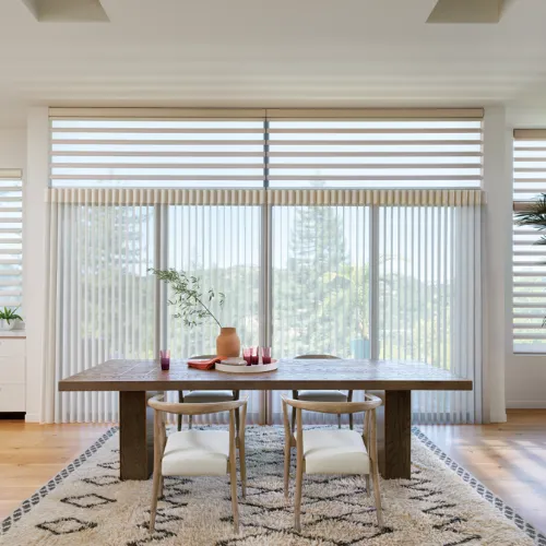 Bright dining room with a wooden table, two light chairs, a vase with branches, and large windows with vertical blinds.