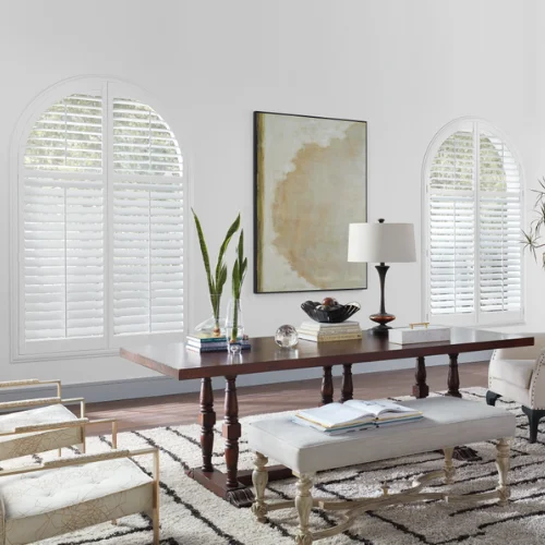 Bright dining/living area featuring a long dark wood table, white benches, and arched windows with blinds over a patterned rug.