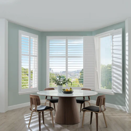 Bright dining nook with a round white table, wooden chairs, and a fruit bowl centerpiece by large bay windows with white shutters open to greenery.