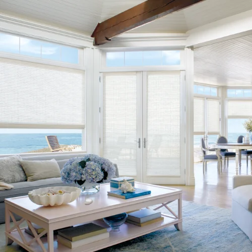 Bright coastal living room with large windows and an ocean view; white furniture, blue hydrangea centerpiece, and light blue rug.