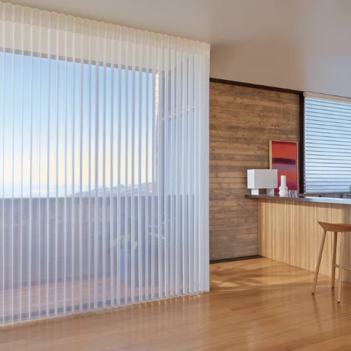 Bright modern living area with sheer vertical blinds, warm wooden floors, and a kitchen island with a stool on the right.