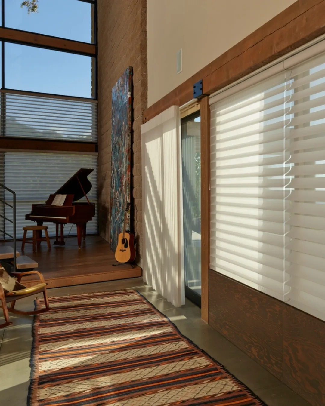 Sunlit music room with a grand piano, an acoustic guitar, bold wall art, and a long striped rug by large windows.