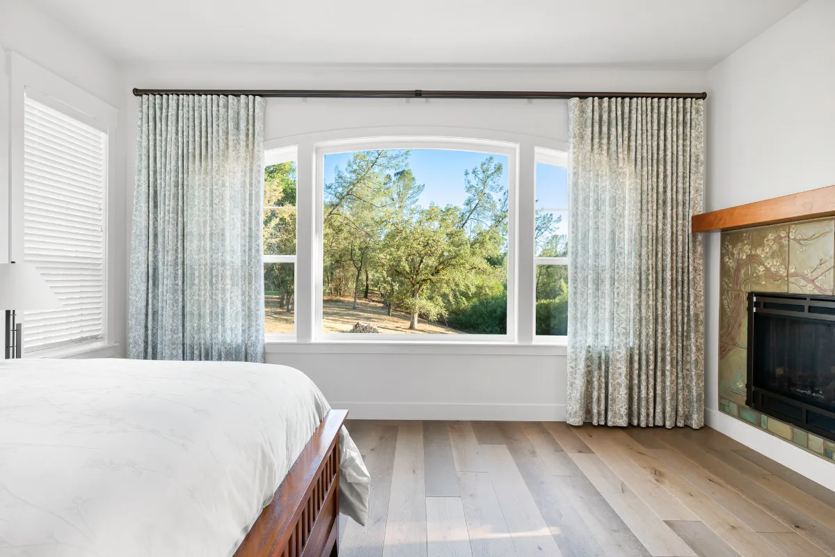 Bright bedroom with a large curved window and blue-gray curtains, trees visible outside, and a wooden bed in the foreground.