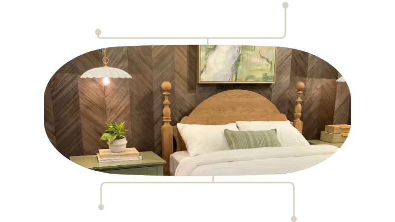 Cozy bedroom with a wooden headboard, white bedding, and a green throw pillow; a nightstand with a plant and books sits to the left, and a painting hangs above the bed.