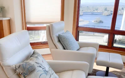 Bright living room with white leather chairs and an ottoman by large bay windows overlooking a lake scene.