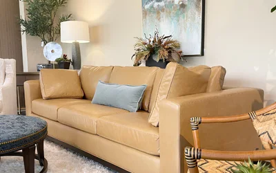 Tan leather sofa with a light blue cushion in a bright living room, with plants, a lamp, and an abstract painting on the wall.