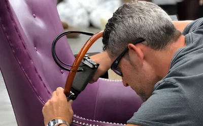 Man wearing safety glasses repairs a purple upholstered chair, using a tool with a brown hose in a workshop.
