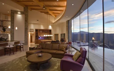 Luxurious hotel lobby with floor-to-ceiling windows offering mountain views, a brown sectional sofa, purple chairs, and a round coffee table near a fireplace and reception area.