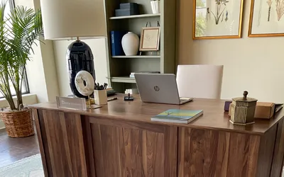 Wooden desk in a home office with a laptop, plant, lamp, bookshelf, and framed botanical art.