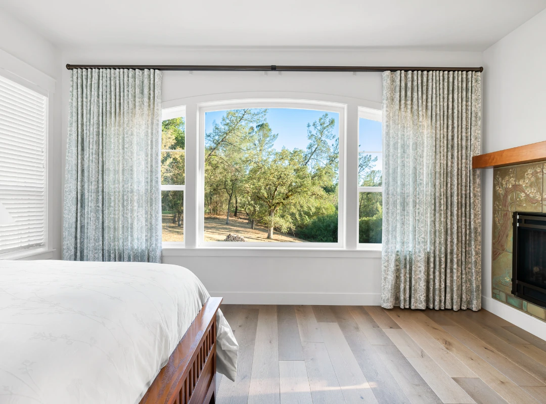 Bright bedroom with a large bay window showing trees outside, white bedding, and a fireplace on the right.