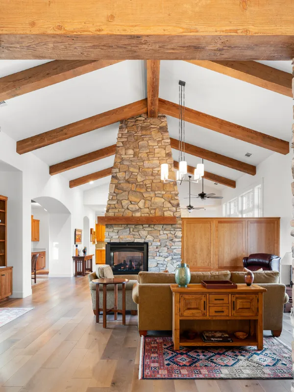 Open living room with a tall stone fireplace reaching the ceiling and exposed wooden beams across a bright, airy ceiling.