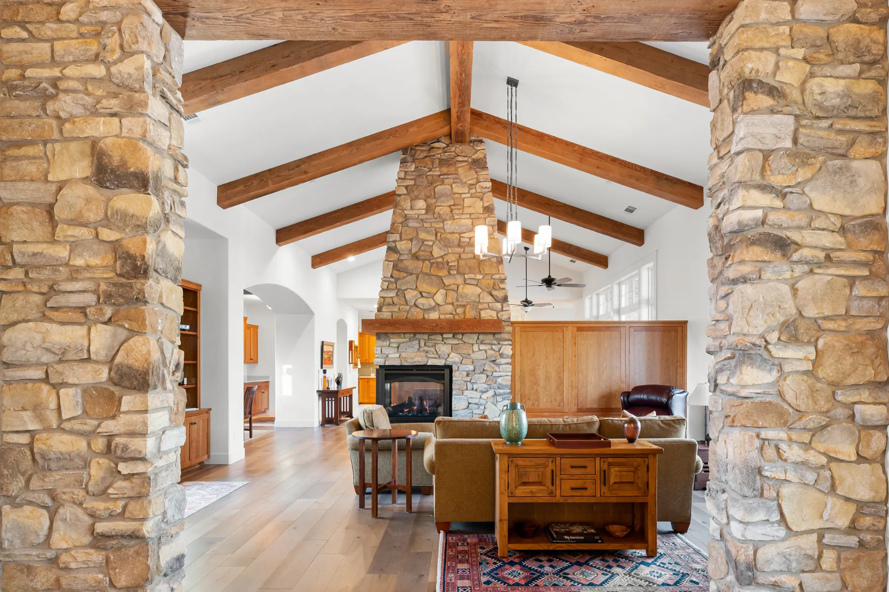 Cozy living room with a tall stone fireplace and exposed wooden beams, flanked by stone pillars and a warm seating area.